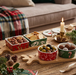 A styled holiday tabletop scene with several Christmas-themed ceramic bowls filled with nuts, crackers, olives, and chocolates, arranged on wooden trays beside a lit candle, pinecones, holly, and a sofa in the background.