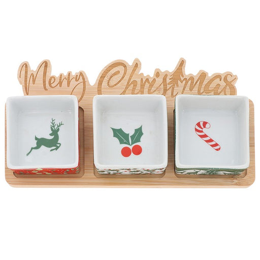 Three small square Christmas-themed dishes on a wooden tray with a wooden sign reading 'Merry Christmas' behind them. The left dish shows a green reindeer, the center dish shows holly with red berries, and the right dish shows a candy cane.