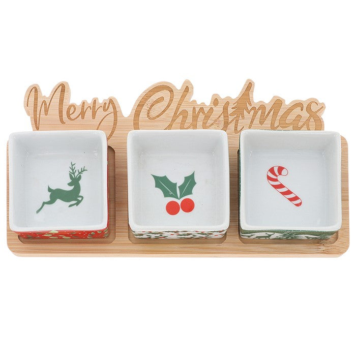 Three small square Christmas-themed dishes on a wooden tray with a wooden sign reading 'Merry Christmas' behind them. The left dish shows a green reindeer, the center dish shows holly with red berries, and the right dish shows a candy cane.