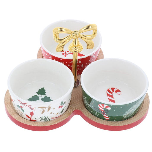 Three Christmas-themed ceramic bowls on a matching wooden and red base, with a gold bow-shaped stand in the center. The bowls feature festive holiday prints including holly, candy canes, pine trees, and red berries.