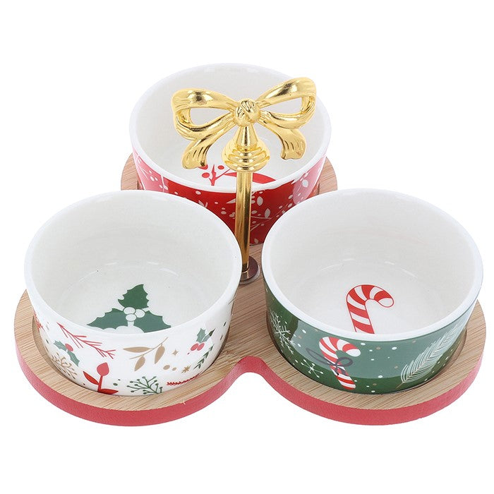 Three Christmas-themed ceramic bowls on a matching wooden and red base, with a gold bow-shaped stand in the center. The bowls feature festive holiday prints including holly, candy canes, pine trees, and red berries.